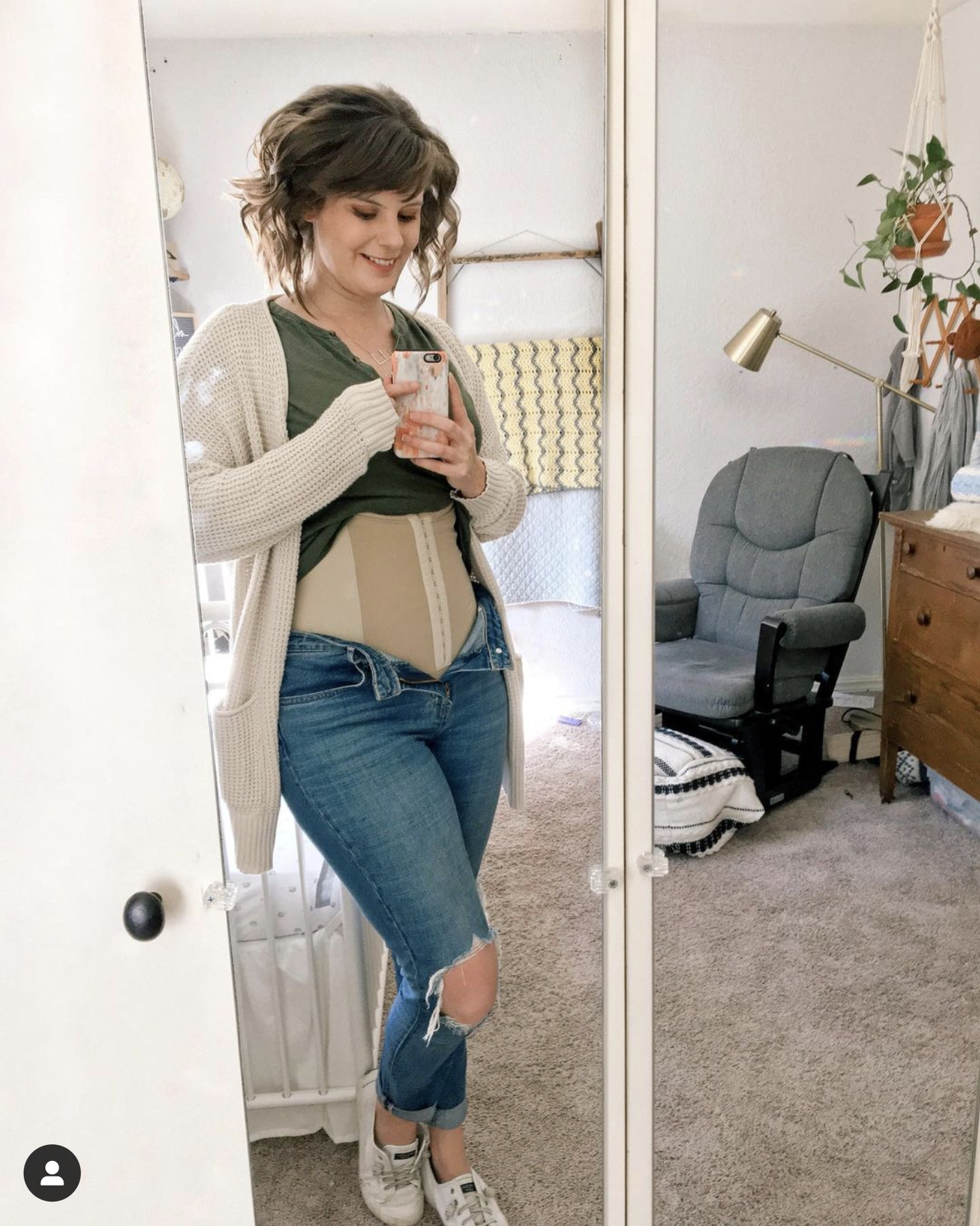 woman standing by crib with a rocking chair in the background. She is wearing jeans. Her shirt is pulled up to show her postpartum wrapping.